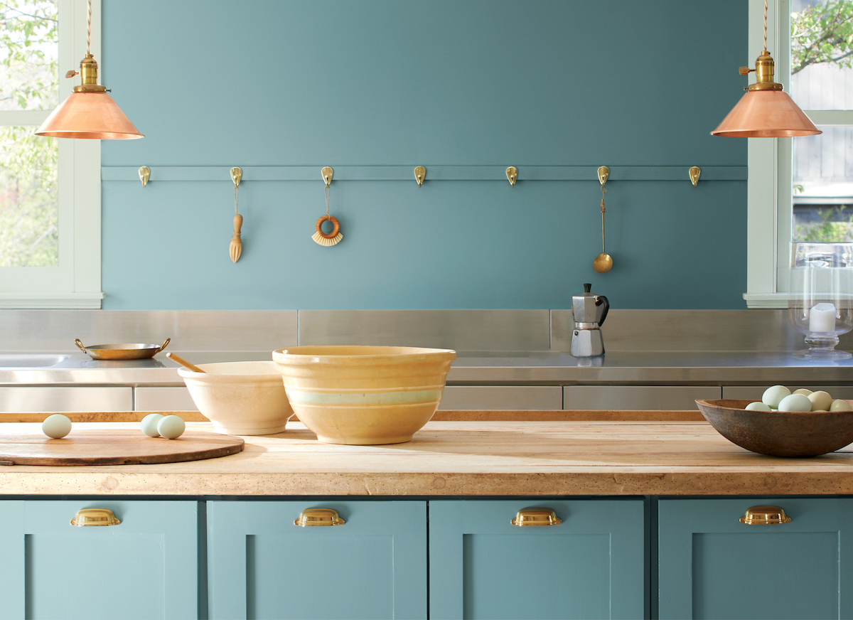 Teal-blue kitchen design featuring warm copper light fixtures, brass accents, and natural wood surfaces.