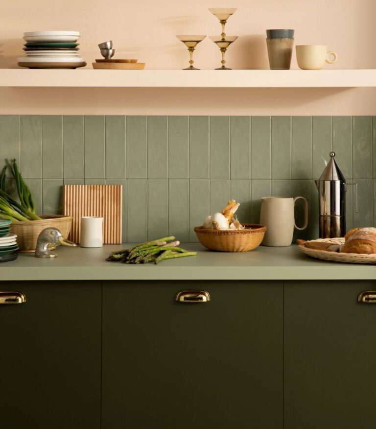 A stylish kitchen with sage green tiled backsplash, dark green cabinets with gold handles, and open shelves displaying neutral-toned dishware. Fresh vegetables, bread, and kitchen essentials add warmth to the earthy color palette.