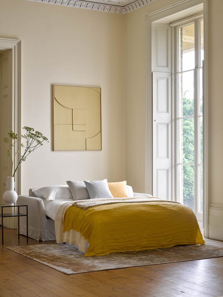 Minimalist bedroom with a neutral color palette, featuring magnolia walls a mustard yellow duvet, soft layered bedding, and a modern abstract wall art piece. Large windows with white shutters allow natural light to brighten the space, highlighting the elegant high ceilings and classic molding details. A simple black side table with a white vase and greenery adds a touch of organic decor.